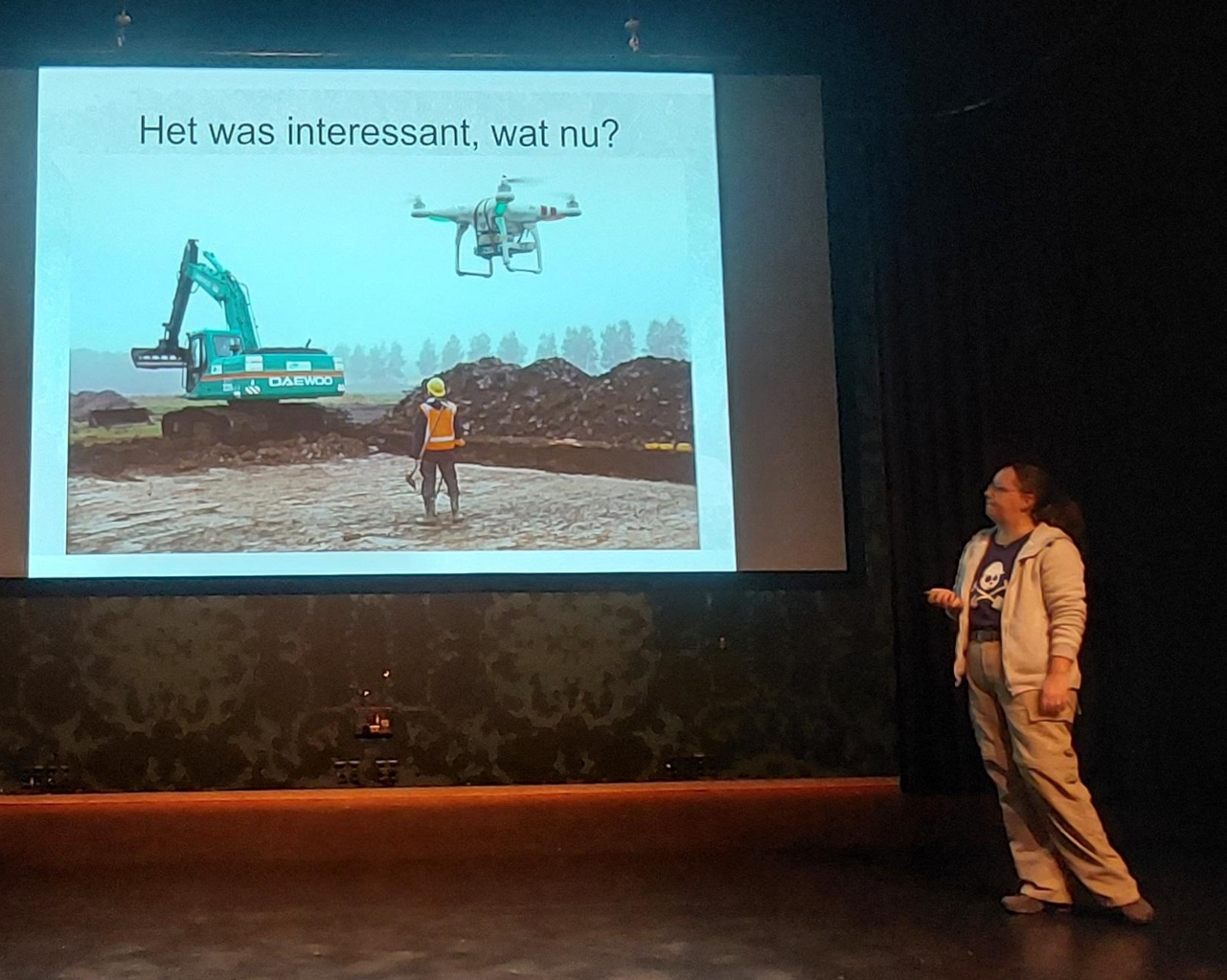 Janneke staat voor een groot projectiescherm met daarop een sheet met een graafmachine, een persoon met oranje hesje en gele helm en op de voorgrond een drone bij een afgevlakt stuk grond. Er naast liggen bergen aarde. Bovenaan de sheet staat: 'Het wass interessant, wat nu?'